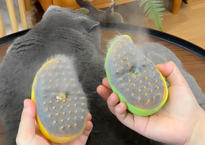 both of the variant of the Steamy Brush(yellow and green) being held upside down showcasing the great amount of cat hair they have collected while emitting steam. A gray cat laying down is visible in the background after it has been massaged